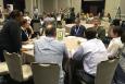 People gather around multiple tables in a large room, some writing on poster boards, discussing science.