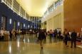 Exhibitors stand with other people at tables on both sides of a long hallway/atrium with high ceilings.