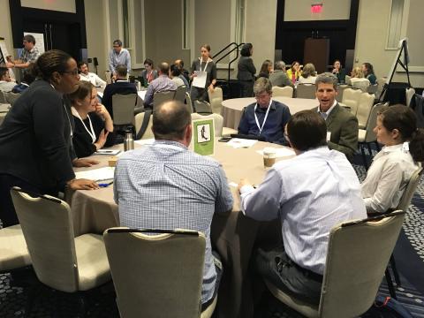 People gather around multiple tables in a large room, some writing on poster boards, discussing science.