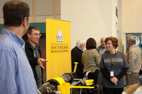 People gather around an exhibitor's booth, with an underwater remotely operated vehicle displayed on a table.