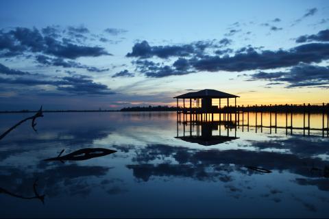 The sun sets across a small bay behind the coastline. A dock and shelter sit above the water in the foreground.