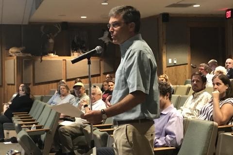 A man stands at a microphone addressing what we can assume is a panel of staff members, with an audience to his back.