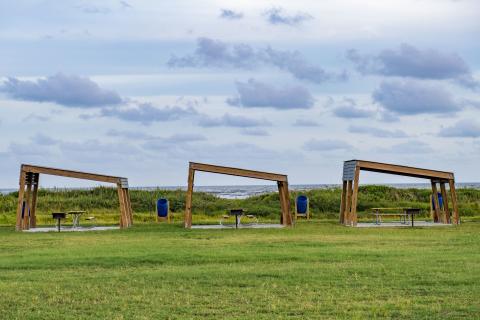 three wooden canopy-like structures sit in a grass field with blue sky in the background