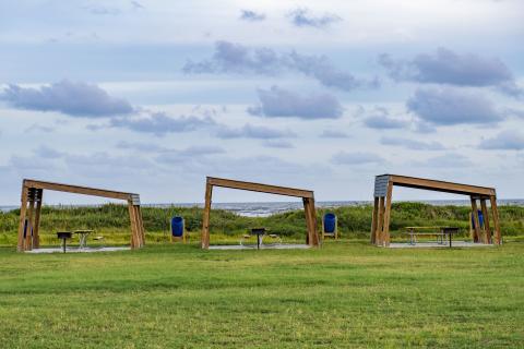 three wooden canopy-like structures sit in a grass field with blue sky in the background
