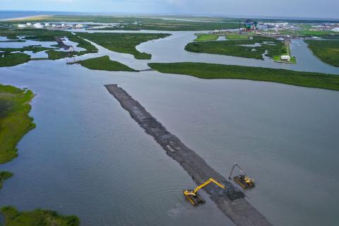 Aerial image of construction equipment moving dirt into a ridge surrounded by marsh and water.