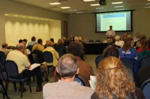 A five-person panel sits at a long table presenting to an audience