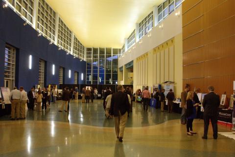 Exhibitors stand with other people at tables on both sides of a long hallway/atrium with high ceilings.