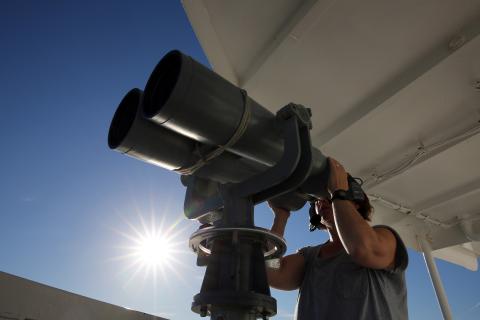 NOAA scientist looks through giant binoculars on a ship, looking for marine mammals.
