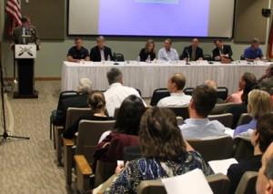 An eight-person panel sits at a long table in front of an audience