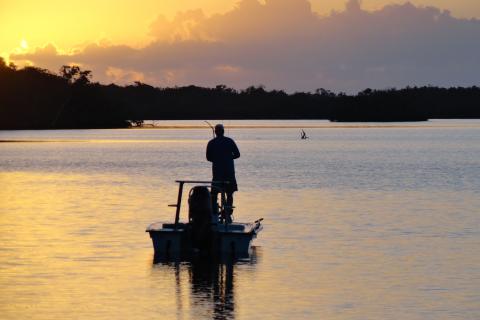 a man stands on a boat in calm water while fishing, with an orange sunset sky with clouds in the background